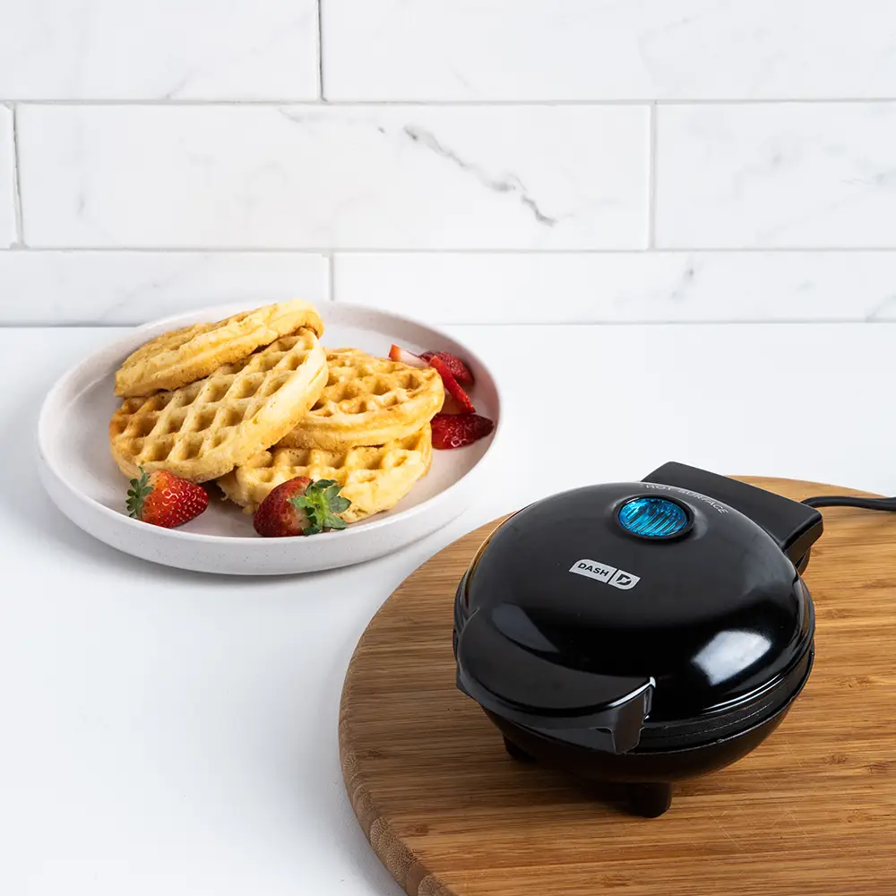 A Dash Mini Waffle Maker on a small countertop, with a single, round, golden-brown waffle on a plate beside it.