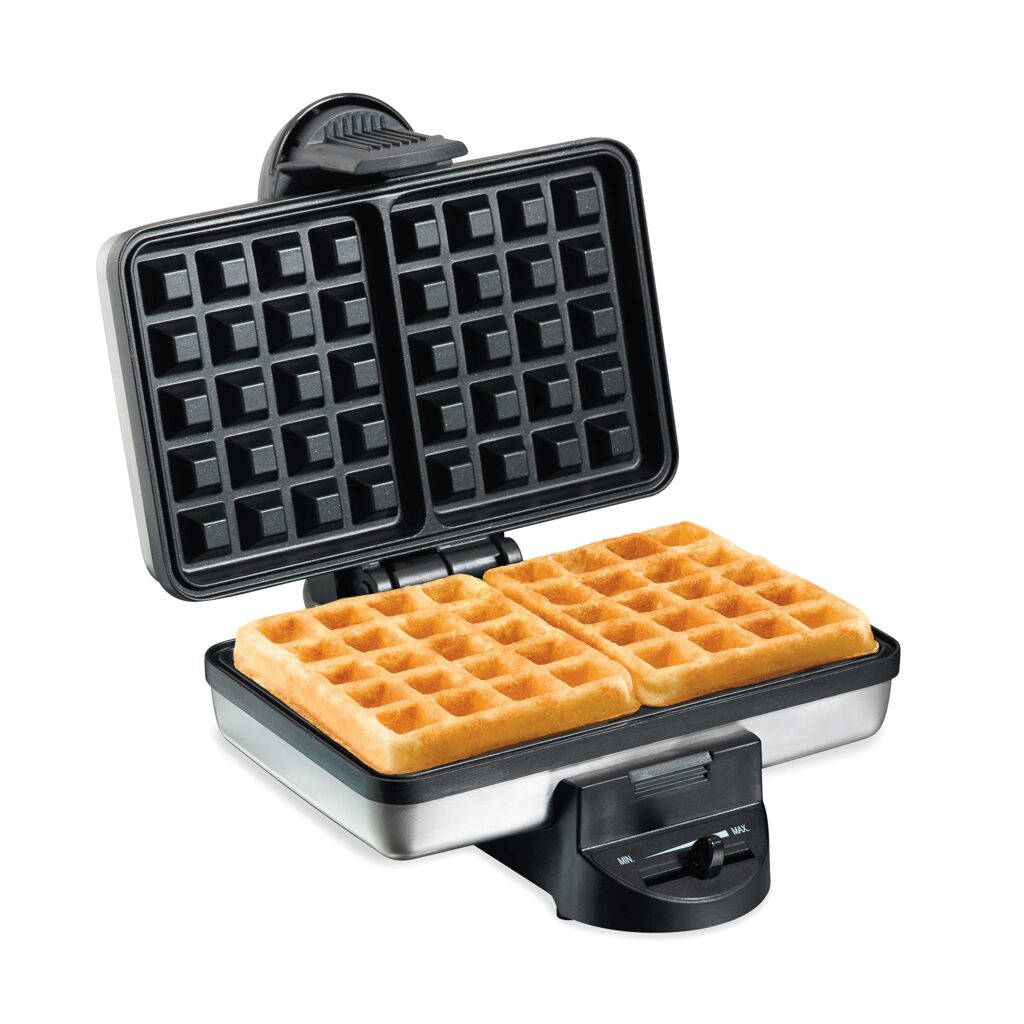 A compact Hamilton Beach Belgian Waffle Maker on a kitchen counter, with a plate of two fluffy Belgian waffles beside it.