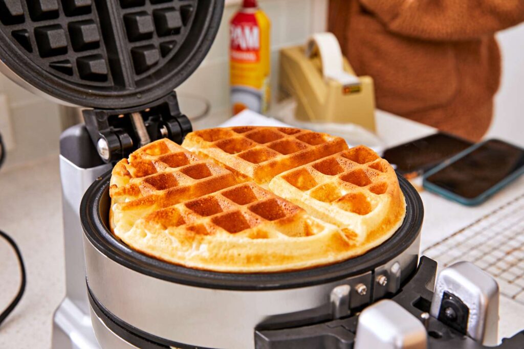 Close-up of a golden-brown waffle cooking inside a waffle maker, with visible crisp edges and steam rising.