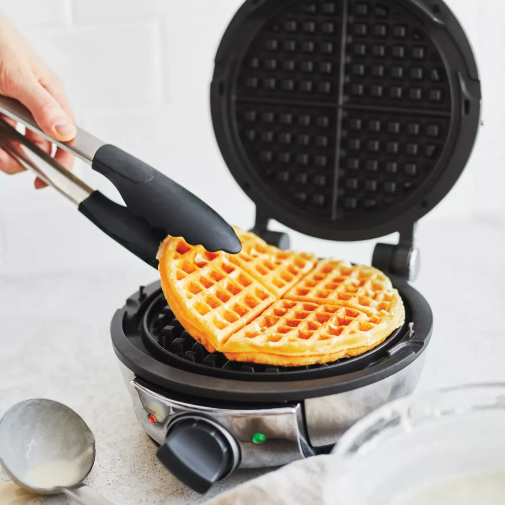 A polished stainless steel All-Clad waffle maker with a round waffle cooking inside, steam gently escaping from the sides.