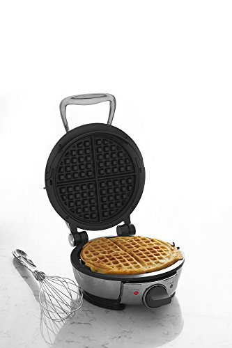 A polished All-Clad Stainless Steel Classic Round Waffle Maker on a marble countertop in a modern kitchen. A waffle is cooking inside, and steam is rising from the appliance's steam-release system.