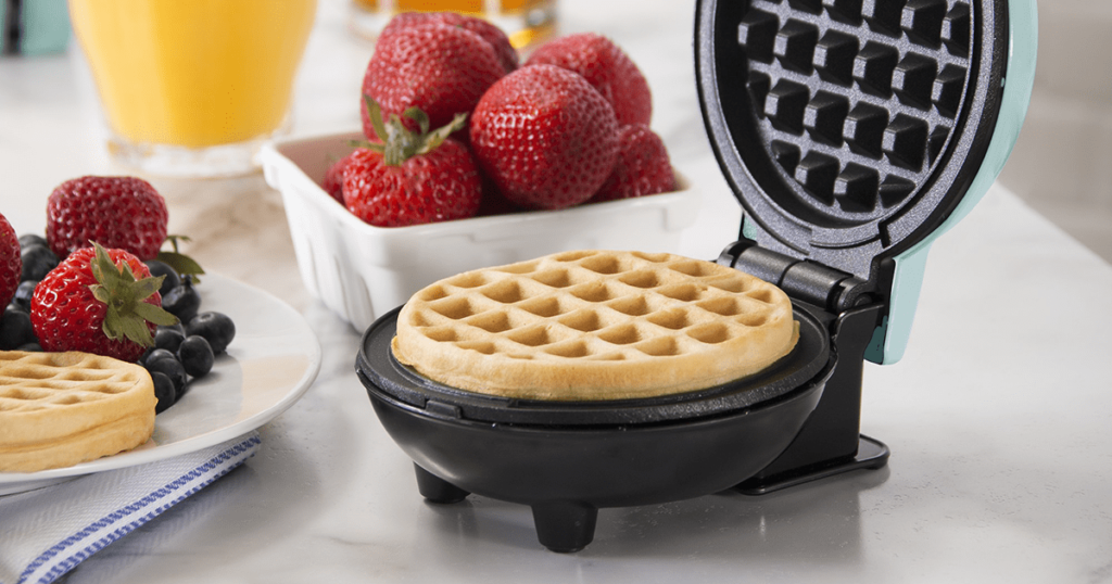 A red Dash Mini Waffle Maker on a small kitchen counter, with a single waffle cooking inside.