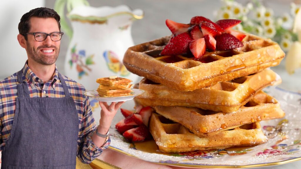 A close-up of a high-end waffle maker with a stainless steel exterior. In the background, a person smiles while eating a perfectly cooked waffle