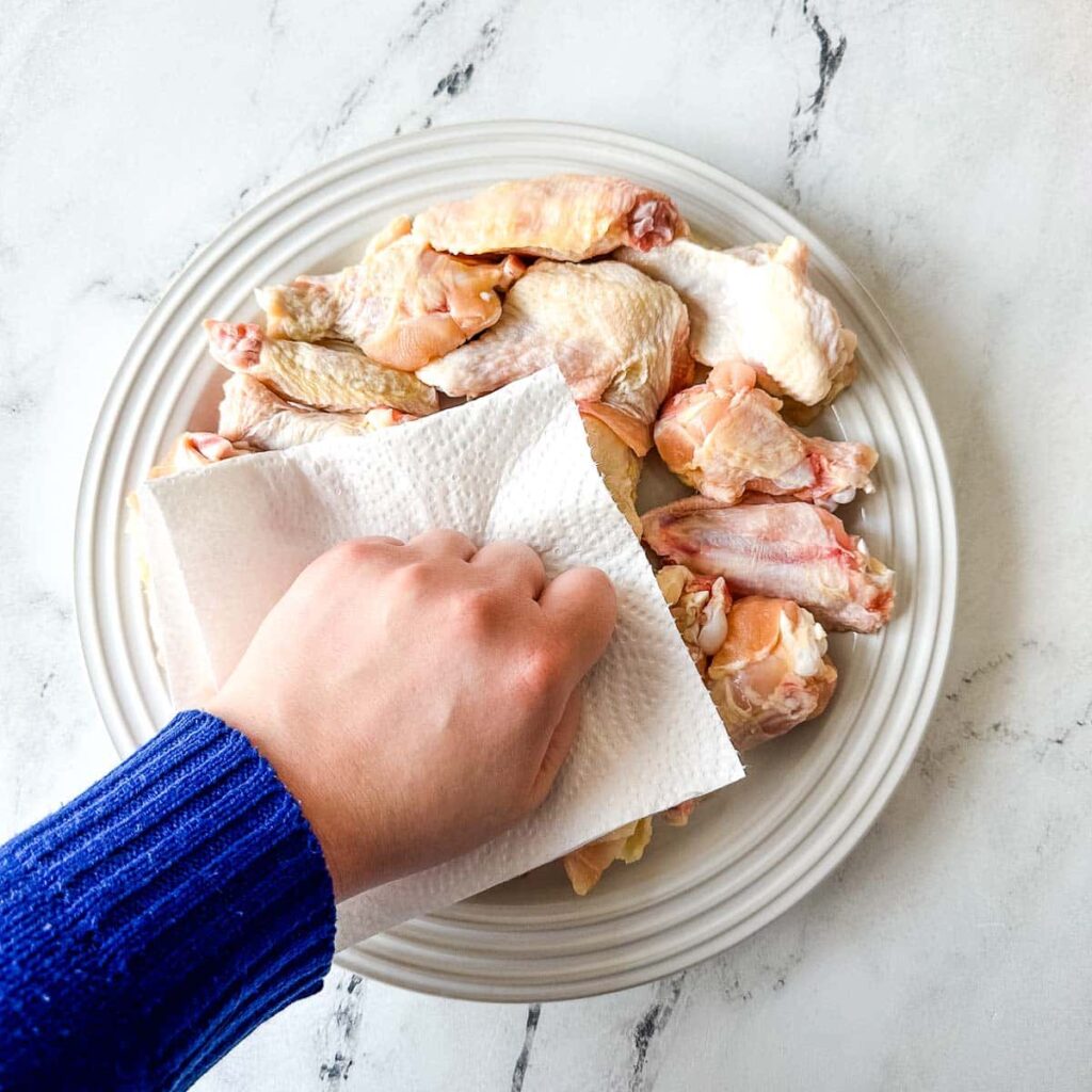 A close-up shot of someone patting chicken wings dry with a paper towel, showing the importance of this step.