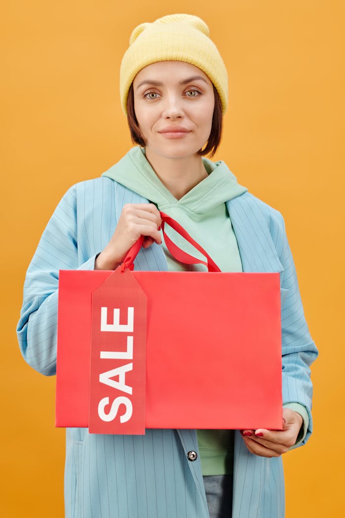 Stylish woman in a blue coat holding a red sale bag against a yellow background, reflecting a modern shopping concept.